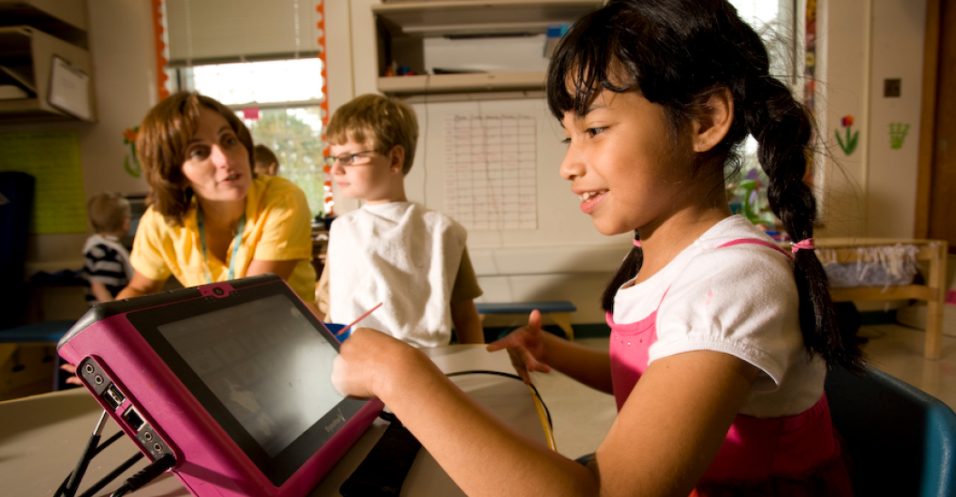 youn girl using AAC device in classroom with teacher