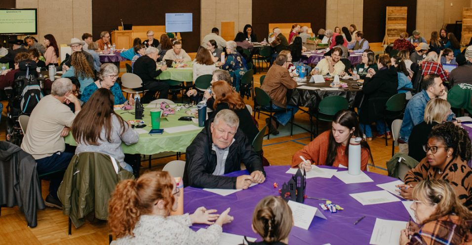 A large community event held in a spacious hall with high ceilings and bright lighting. Multiple round tables covered with colorful tablecloths—purple, green, and other shades—are arranged throughout the room. Each table has papers, notebooks, and water bottles scattered across it. Many people are seated around the tables, engaged in discussions or writing. In the background, there are large wall panels, a scoreboard, and wooden structures stacked near the wall.