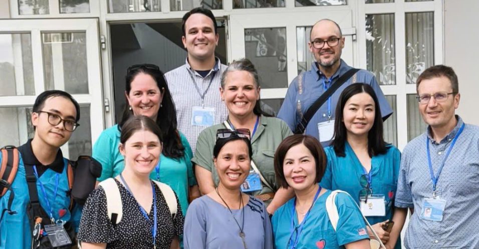 A group of ten adults pose together and smile in front of a building with glass doors